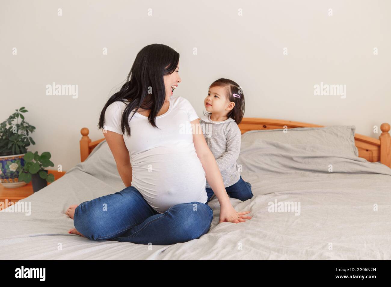 Donna incinta cinese asiatica che gioca con la bambina a letto a casa. Ragazza figlia capretto che si nasconde dietro la mamma. Madre e figlia del bambino che aspettano l'attesa Foto Stock