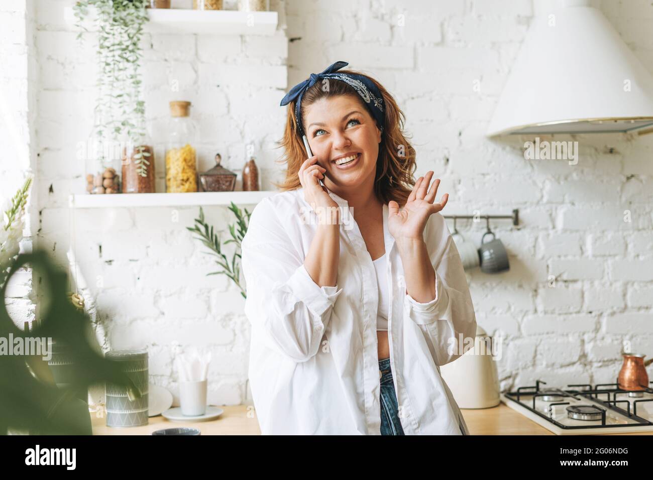 Splendida giovane donna più corpo taglia positivo in jeans blu e camicia bianca che parla sul cellulare in cucina casa Foto Stock