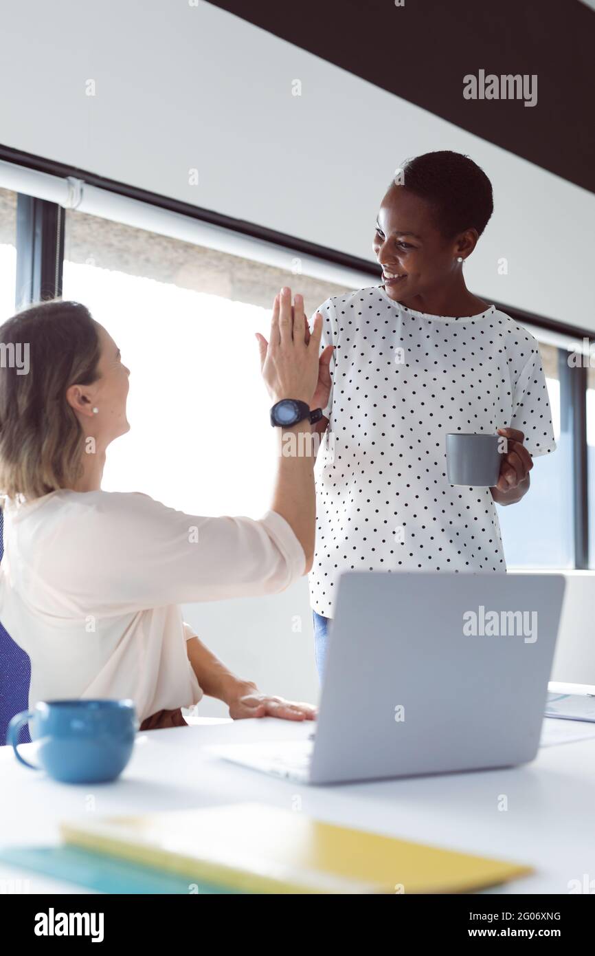 Due diverse donne d'affari alla scrivania, parlando e dando alto cinque Foto Stock