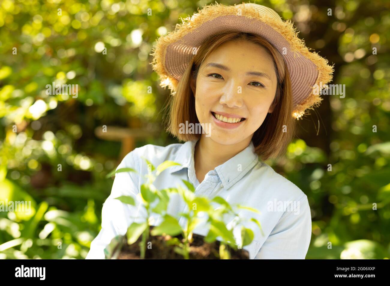 Donna asiatica sorridente che indossa cappello di paglia e tiene la pianta in giardino Foto Stock