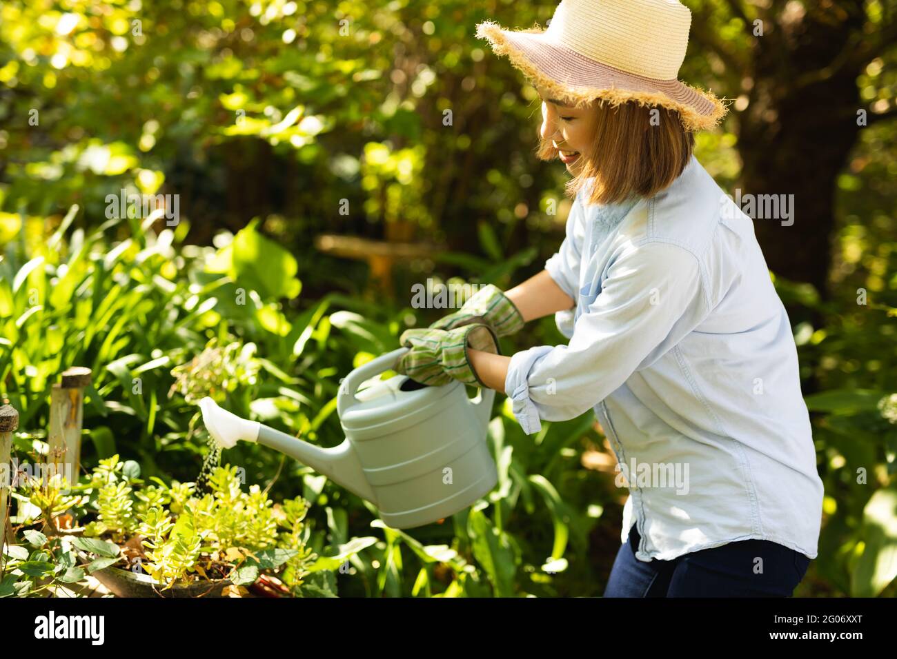 Donna asiatica sorridente che indossa cappello di paglia e che utilizza annaffiatura lattina in giardino Foto Stock