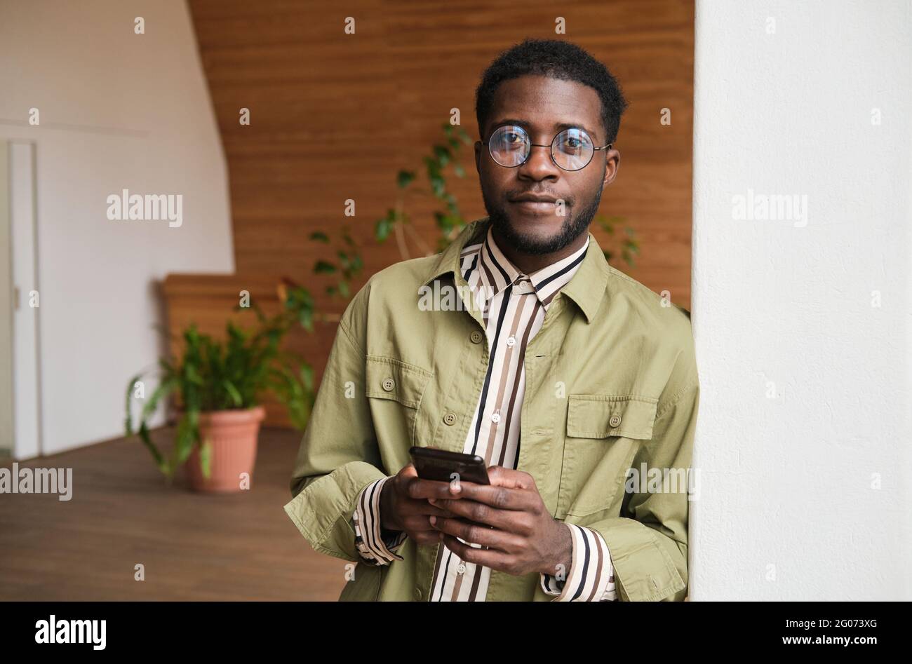 Ritratto di un giovane uomo d'affari africano che guarda la fotocamera e usa il telefono cellulare mentre si trova in ufficio Foto Stock