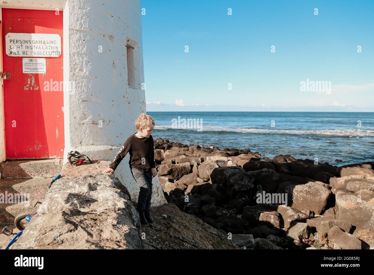 Primo piano di un ragazzo caucasico su una spiaggia oceanica con grandi rocce e un edificio Foto Stock