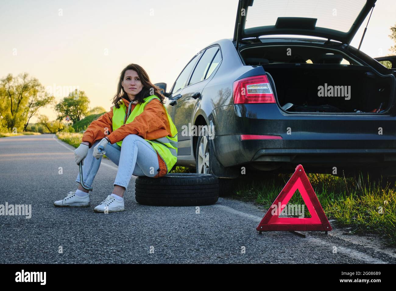 La ragazza cambia la ruota danneggiata della vettura. Giubbotto verde. Ruota di scorta. Incidente stradale. Viaggio in auto. Foto Stock