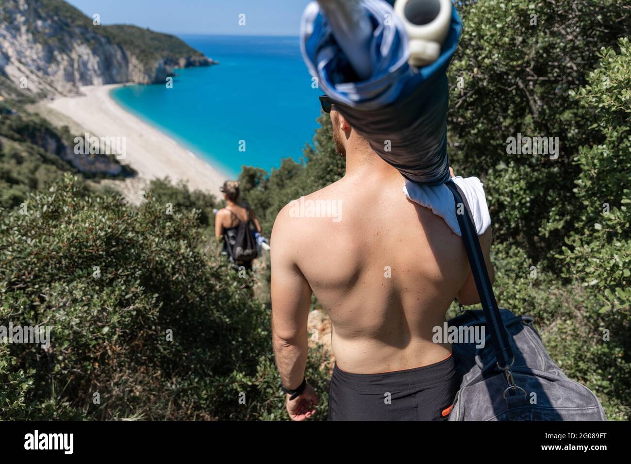 Giovane coppia che scende alla bellissima spiaggia sabbiosa di Milos, portando con sé un ombrellone e altri accessori da spiaggia Foto Stock