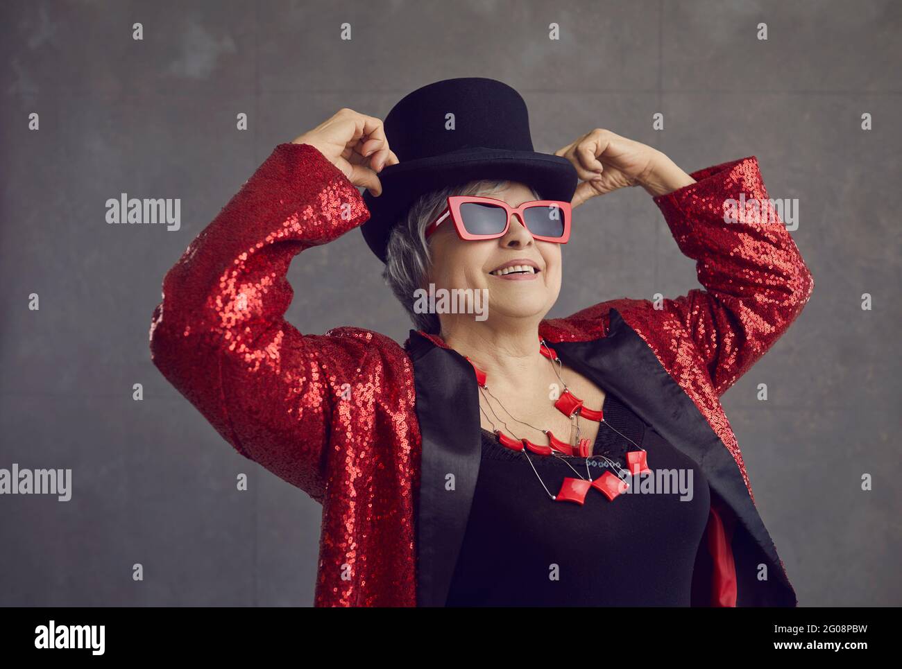 Felice bella donna anziana in top Hat, occhiali di stile e giacca bling in posa in studio Foto Stock
