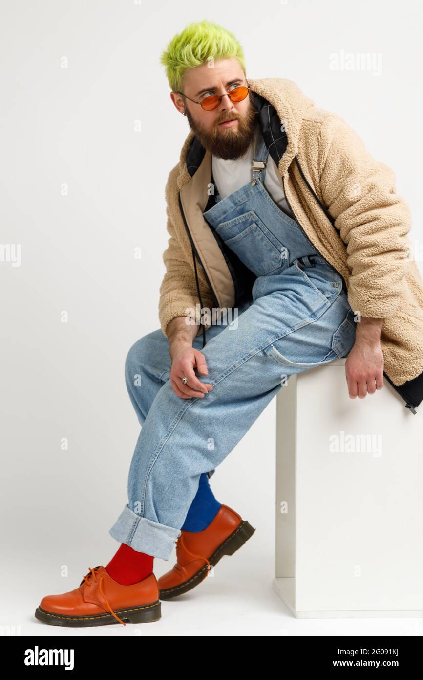 Giovane uomo con barba e capelli verdi alla moda che indossa abiti e scarpe  eleganti, seduto su un cubo bianco e guardando lontano con espressione  penosa. Interno Foto stock - Alamy