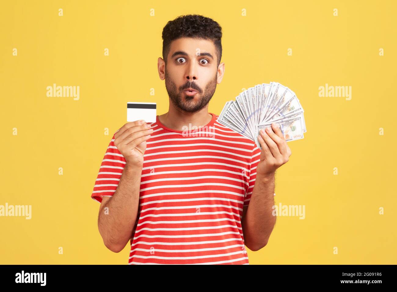 Scioccato uomo bearded in T-shirt a righe tenendo e mostrando il dollaro in contanti e carta di credito, sorpreso con cashback, cambio di valuta. Riprese in studio al coperto Foto Stock