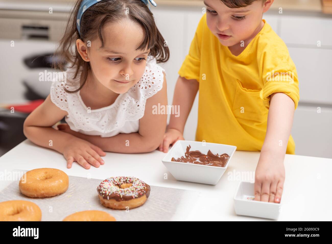 Bambini che decorano ciambelle con spruzzi colorati in cucina a casa Foto Stock