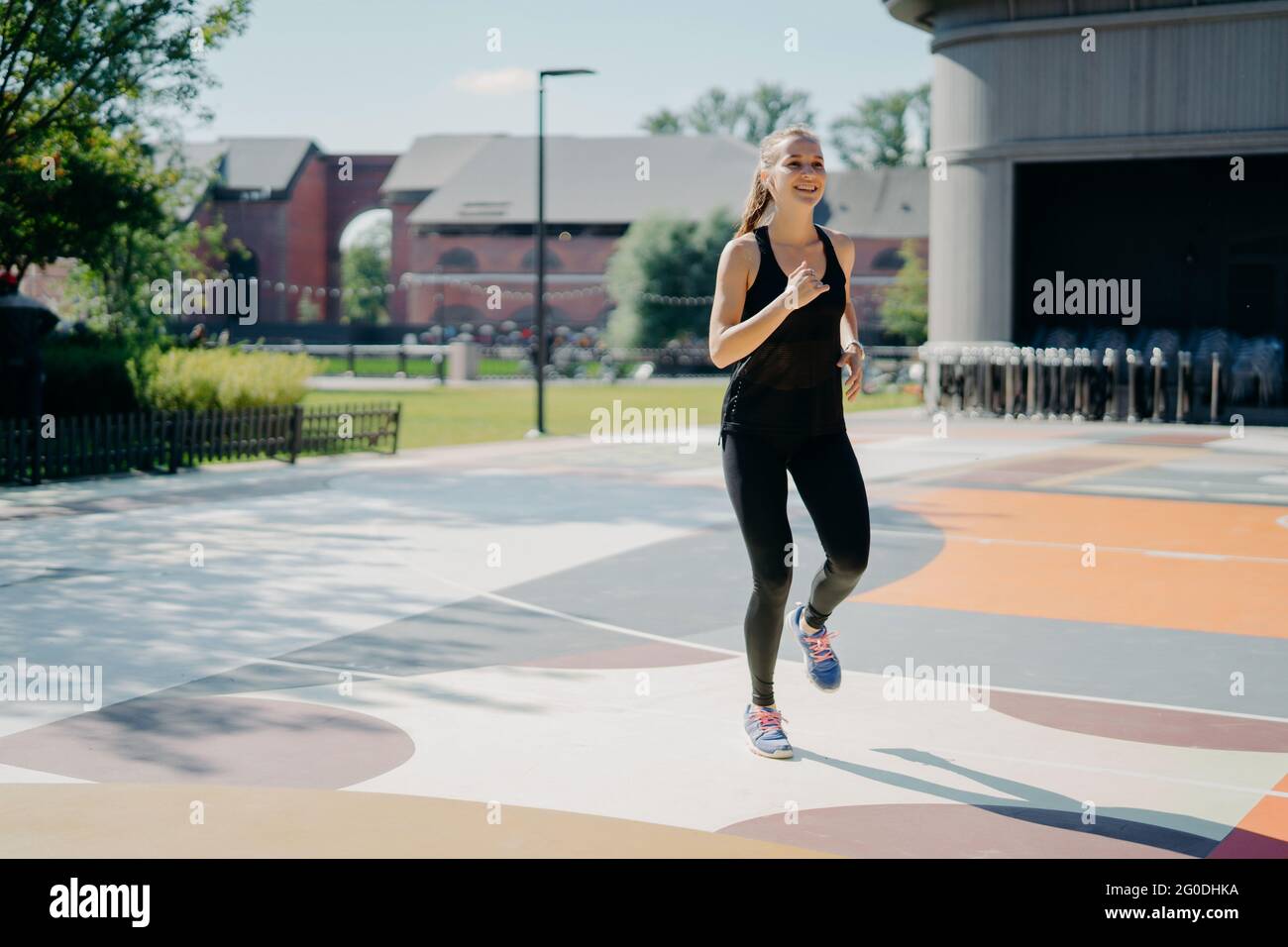 La donna allegra corre attivamente sullo stadio vestito di abiti sportivi neri gode di attività fisiche all'aperto durante la giornata estiva essendo piena di energia Foto Stock
