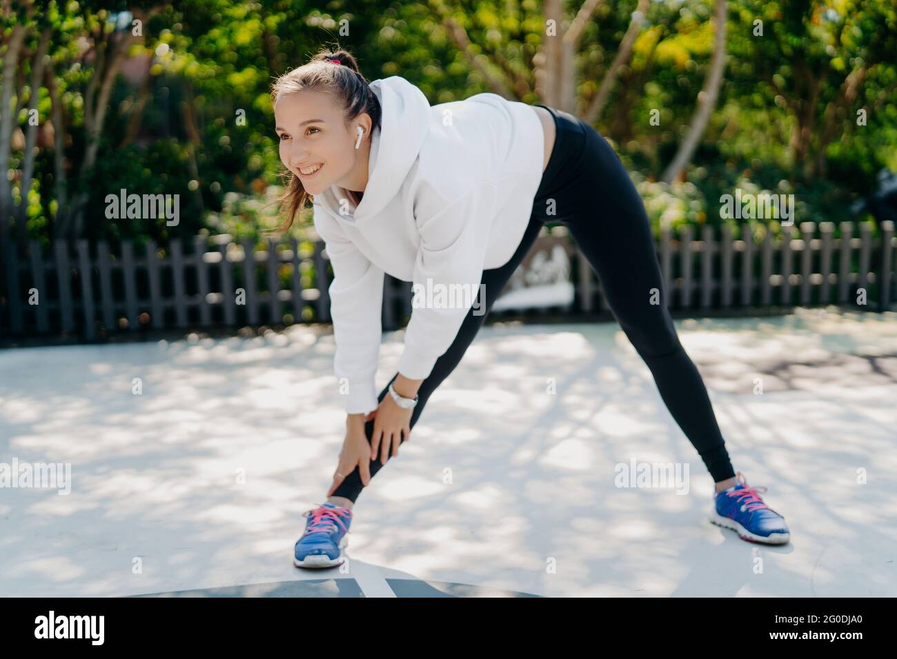 La donna vivace sorride gladfully concentrata in lontananza si appoggia alla gamba vestita di abbigliamento sportivo treni all'aperto essendo motivata ascolta la musica in Foto Stock