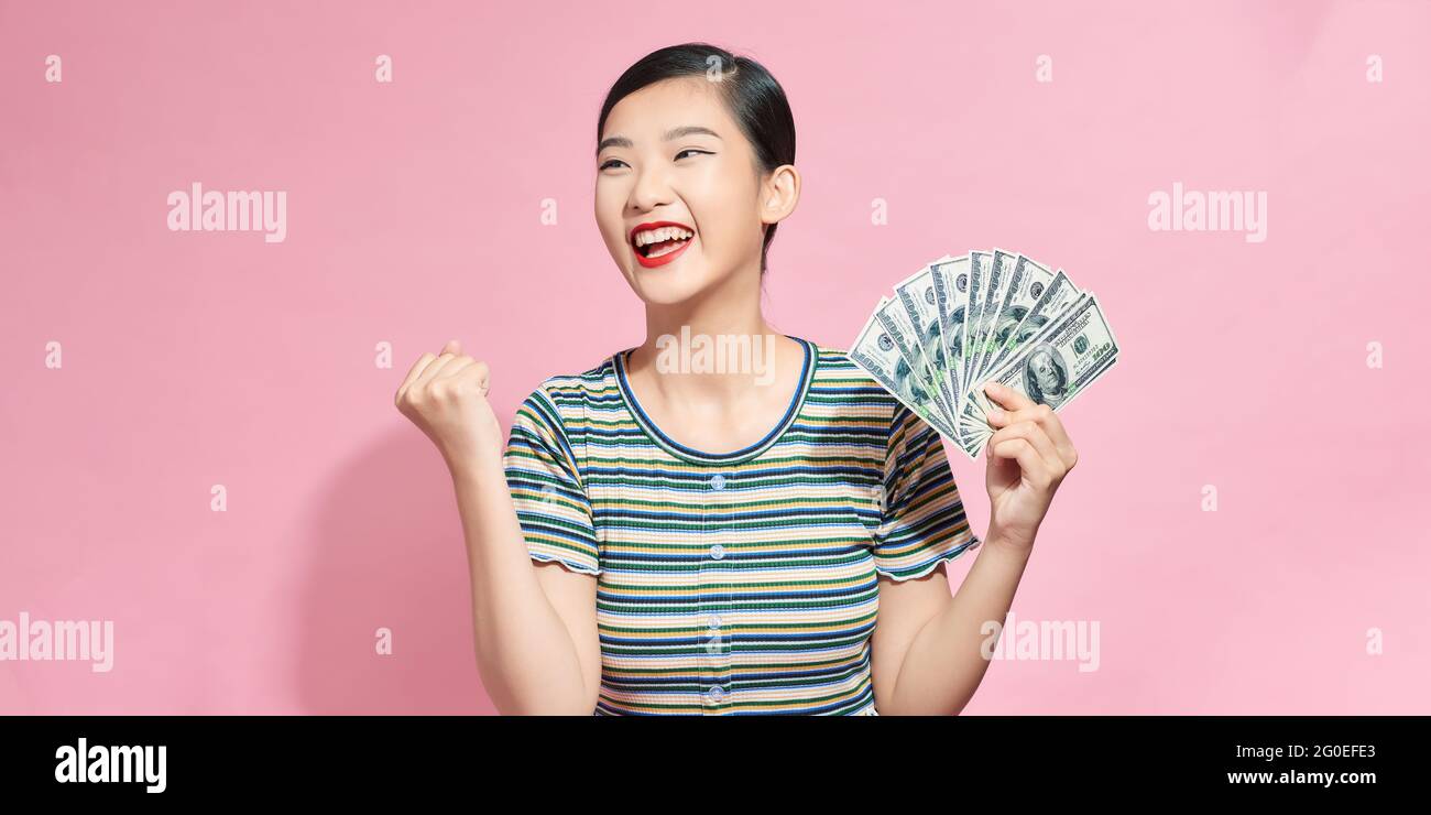 Immagine di una donna fortunata che tiene fan di 100 dollari bollette essere eccitato per vincere il premio in denaro, esprimendo la vittoria e la ricchezza sul muro rosa Foto Stock