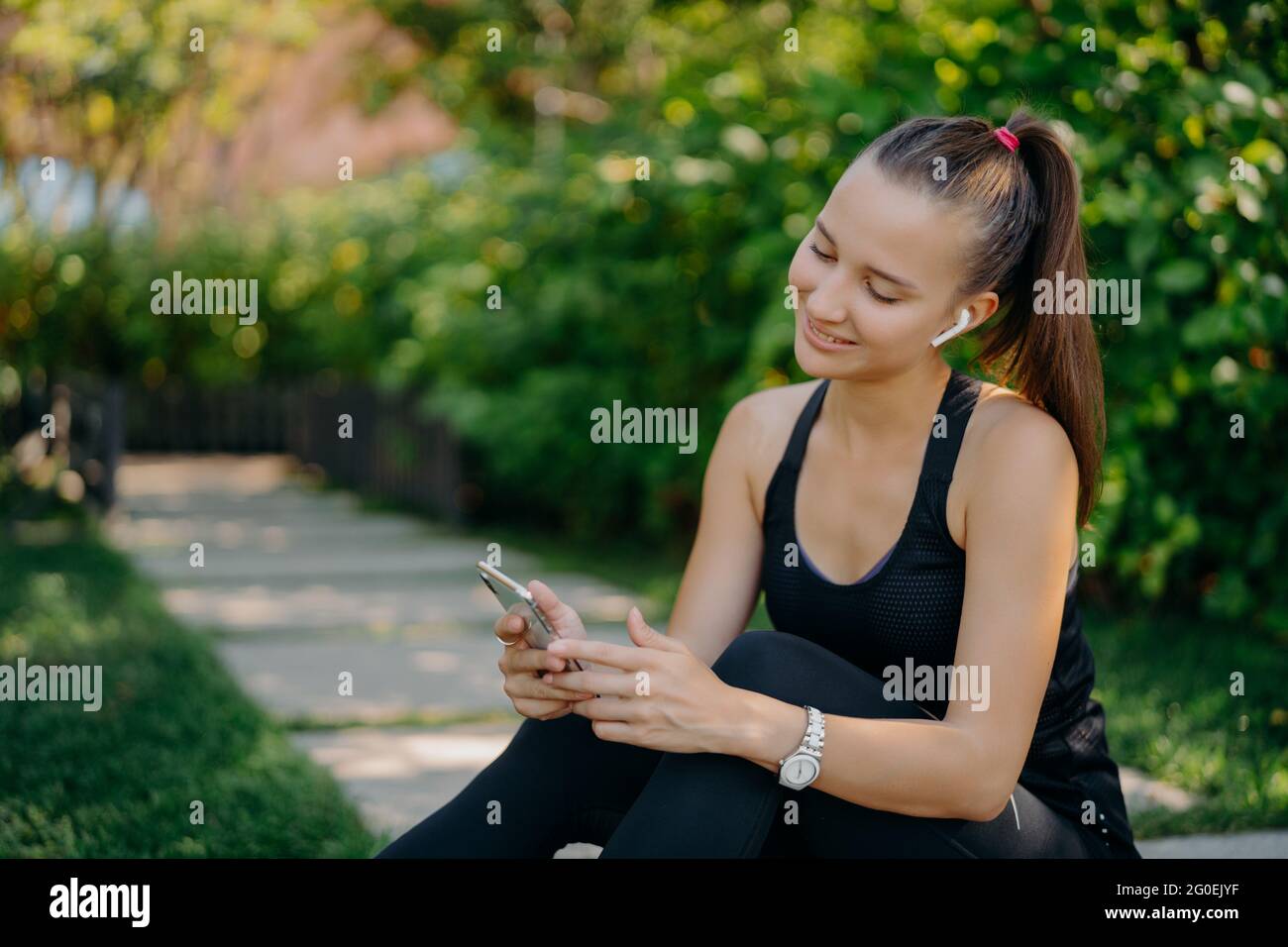 Donna felice con coda pony indossa un abbigliamento sportivo concentrato al dispositivo smartphone ascolta la musica in wireless auricolari navigare internet ha all'aperto Foto Stock