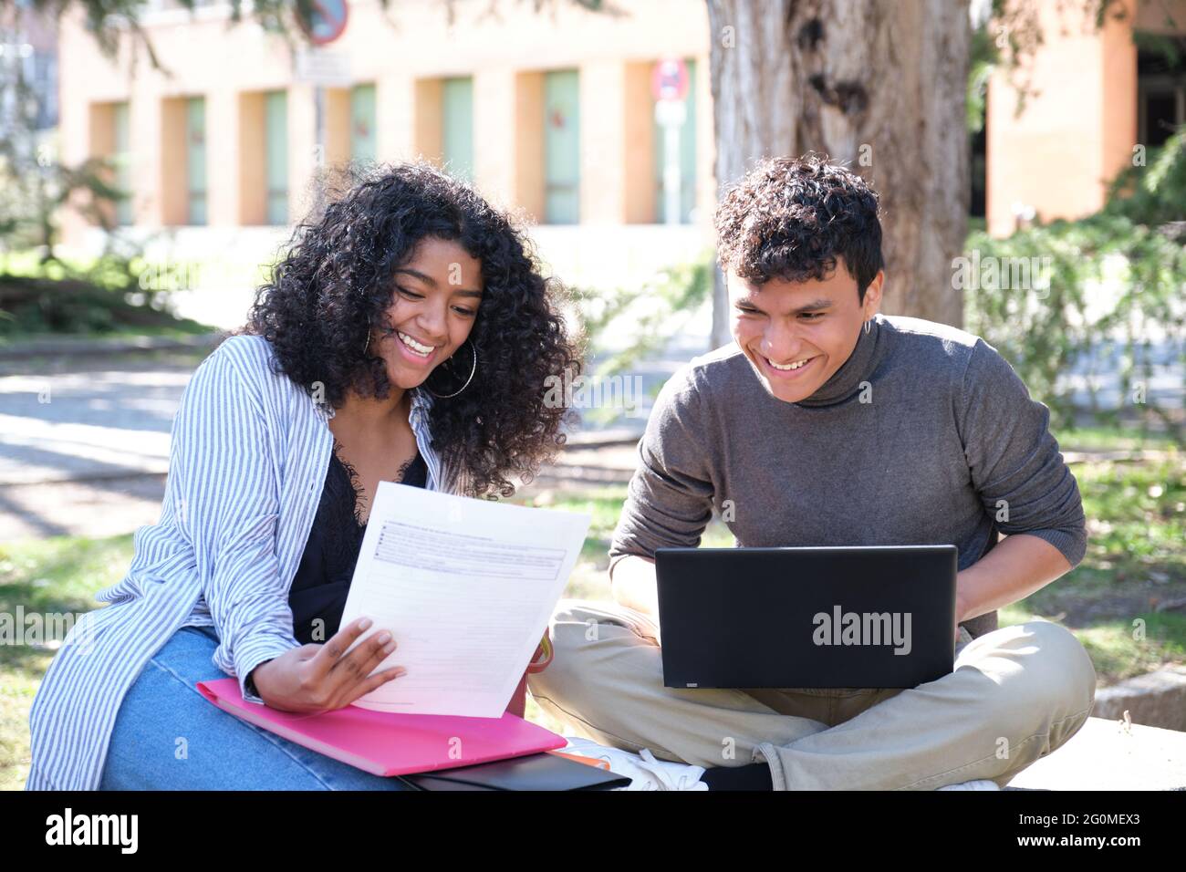 Due studenti latini ridendo studiare insieme seduti su una panchina all'aperto. Vita universitaria. Foto Stock