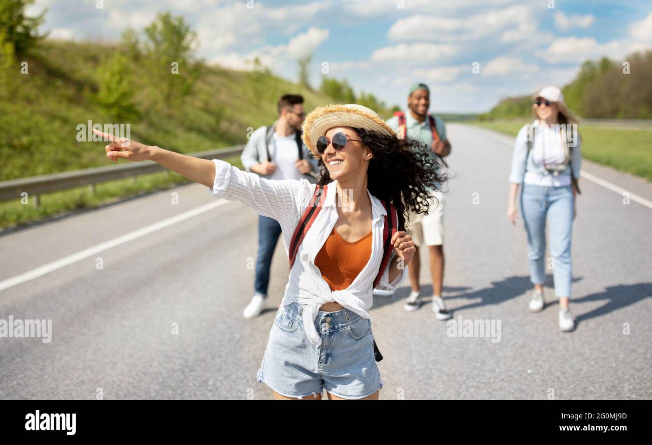 Giovane donna caucasica con i suoi amici multirazziali hitchhiking in autostrada, viaggiando in autostop, all'aperto Foto Stock