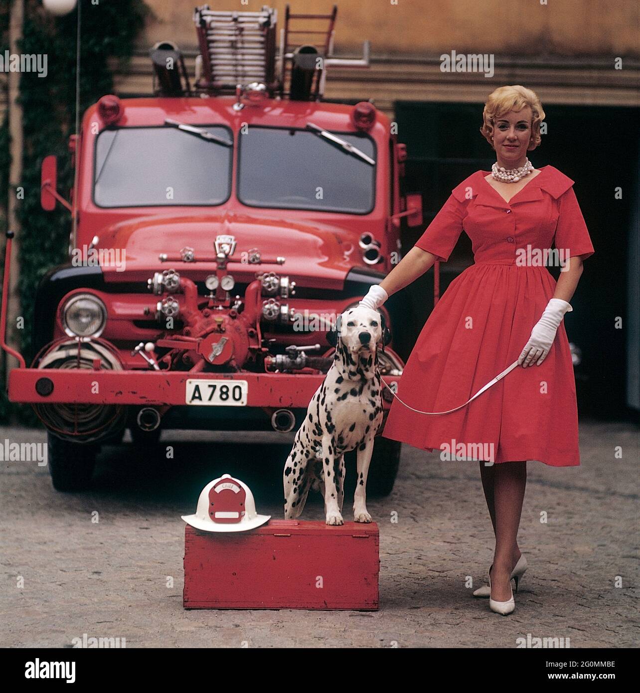 Alla moda negli anni '50. Una giovane donna in un abito rosso, guanti bianchi e scarpe bianche è in piedi di fronte a un camion da fuoco Ford insieme a un cane dalmata con un casco capo fuoco americano. Il dalmatier è tradizionalmente una mascotte della brigata del fuoco. Svezia 1958 rif CV68-11 Foto Stock