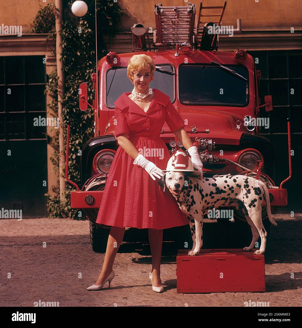 Alla moda negli anni '50. Una giovane donna in un abito rosso, guanti  bianchi e scarpe bianche è in piedi di fronte a un camion da fuoco Ford  insieme a un cane