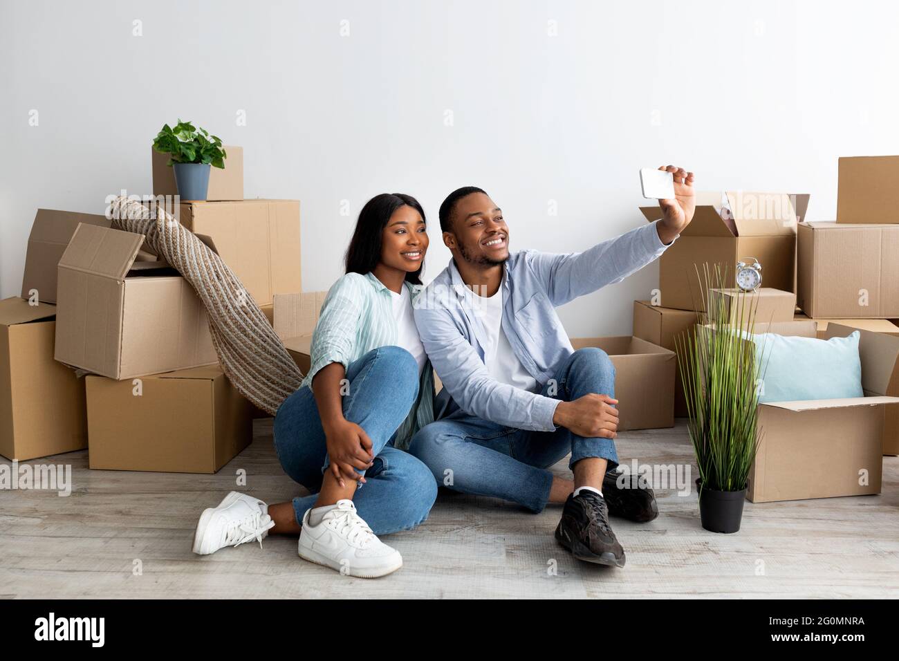 Felice coppia nera che si sposta a casa propria e prendendo selfie tra scatole di cartone, spazio libero Foto Stock