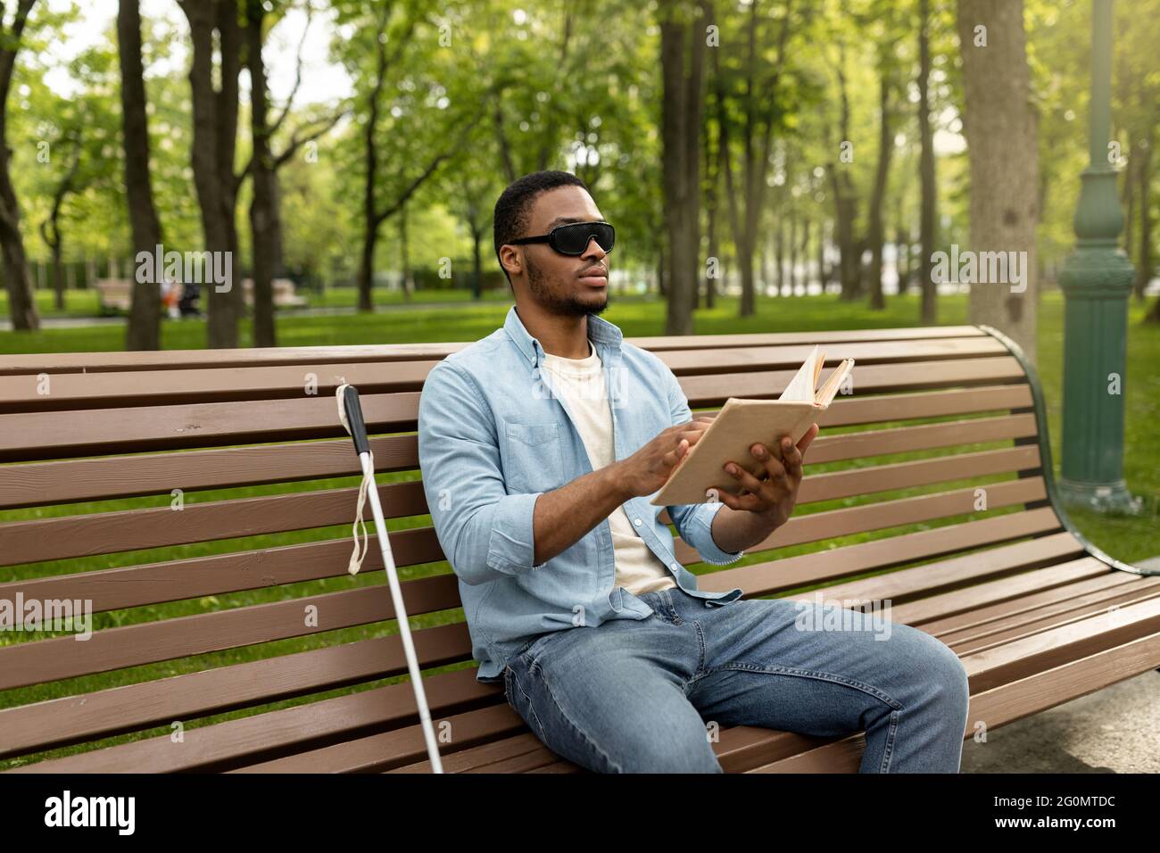 Giovane uomo nero per non vedenti seduto sulla panchina nel parco cittadino, leggendo il libro Braille all'aperto Foto Stock