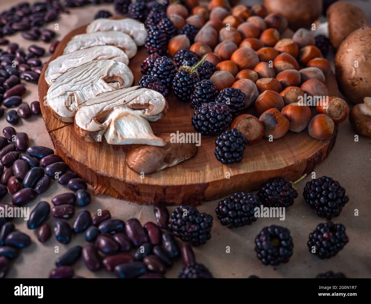 Una sana selezione di cibo autunnale: Funghi bianchi a fette, nocciole sgusciate, fagioli porpora, more e piccolo ramo di abete su una fetta di legno. Foto Stock