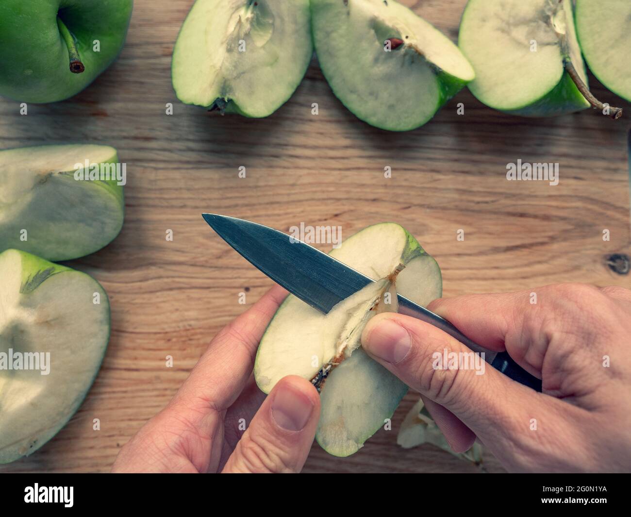 Mano con coltello che taglia un nucleo di mela su un tagliere di legno, freschi quarti di mela verde succosa sparsi sullo sfondo. Foto Stock