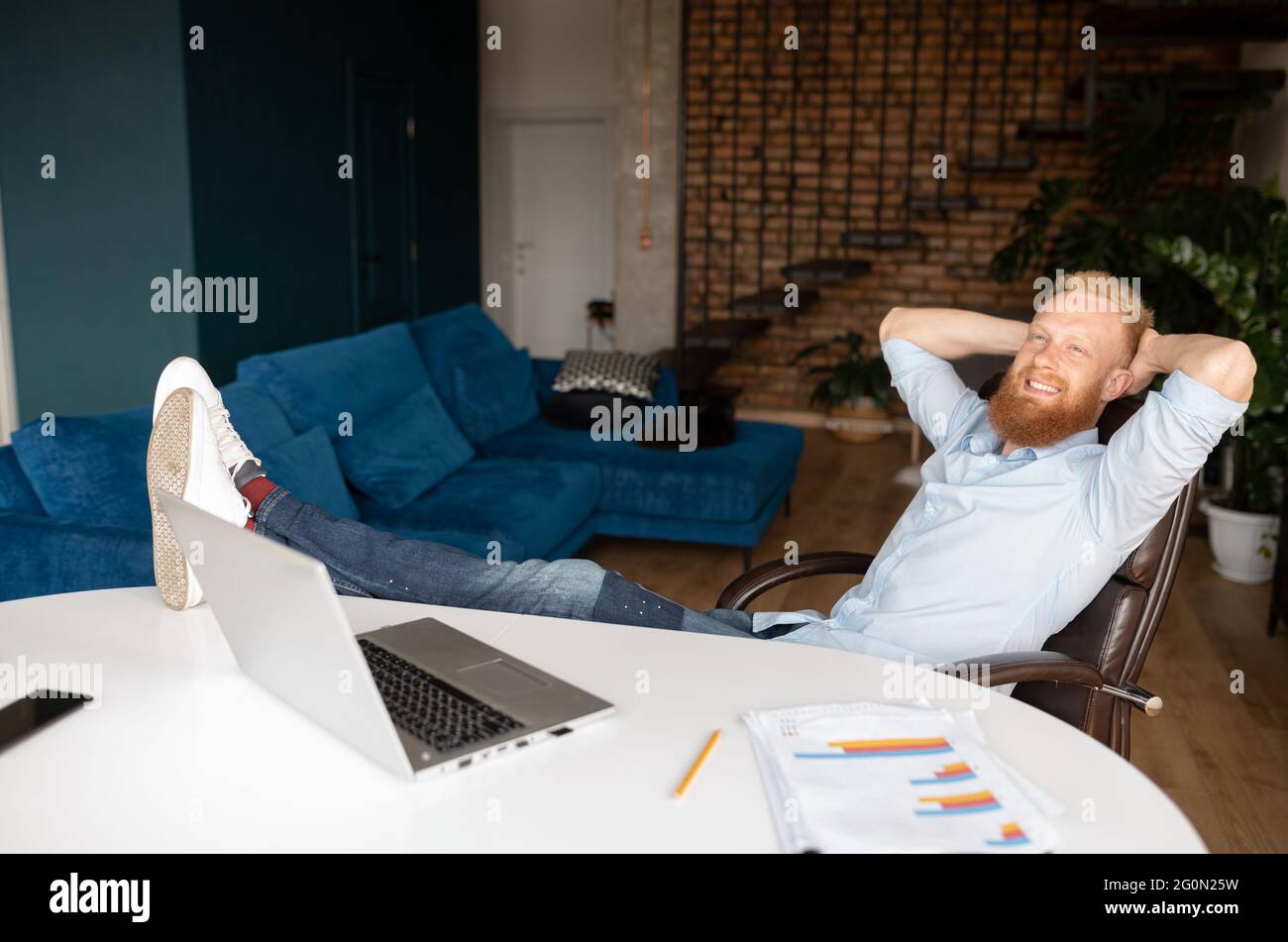 Un giovane imprenditore rosso di successo riposa nell'ufficio domestico con i suoi piedi sulla scrivania, appoggiato sulla sedia con le mani dietro la testa, sorridente uomo che sogna daydreaming di fronte al portatile Foto Stock