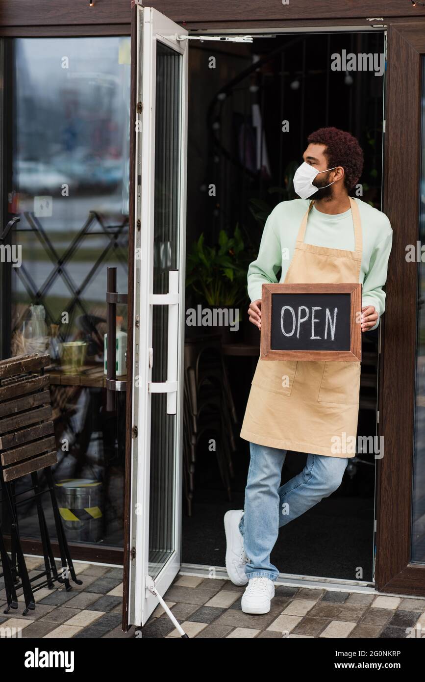 Barista afroamericano in maschera medica che tiene la lavagna con le scritte aperte nel caffè Foto Stock