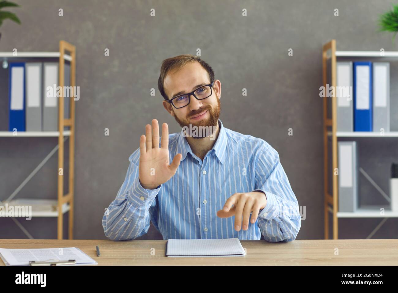 Felice lavoratore in ufficio in occhiali seduti alla scrivania, sorridendo e muovendo la mano alla macchina fotografica Foto Stock