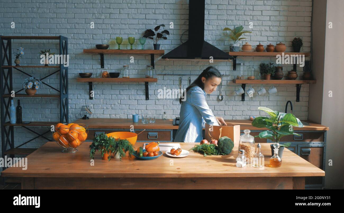 Una bella giovane donna di buon umore sta smistando le verdure da una borsa e metterle sulla tavola della cucina.housewife giovane e felice stabilisce gli acquisti Foto Stock