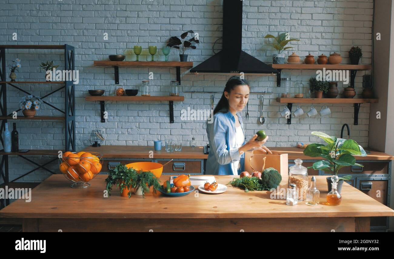Una bella giovane donna di buon umore sta smistando le verdure da una borsa e metterle sulla tavola della cucina.housewife giovane e felice stabilisce gli acquisti Foto Stock