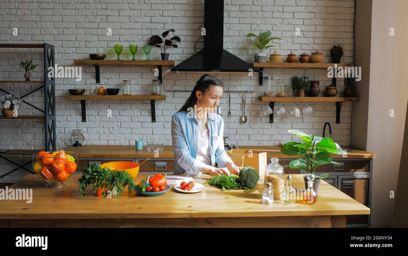 Una bella giovane donna di buon umore sta smistando le verdure da una borsa e mettendole sul tavolo della cucina. Foto Stock