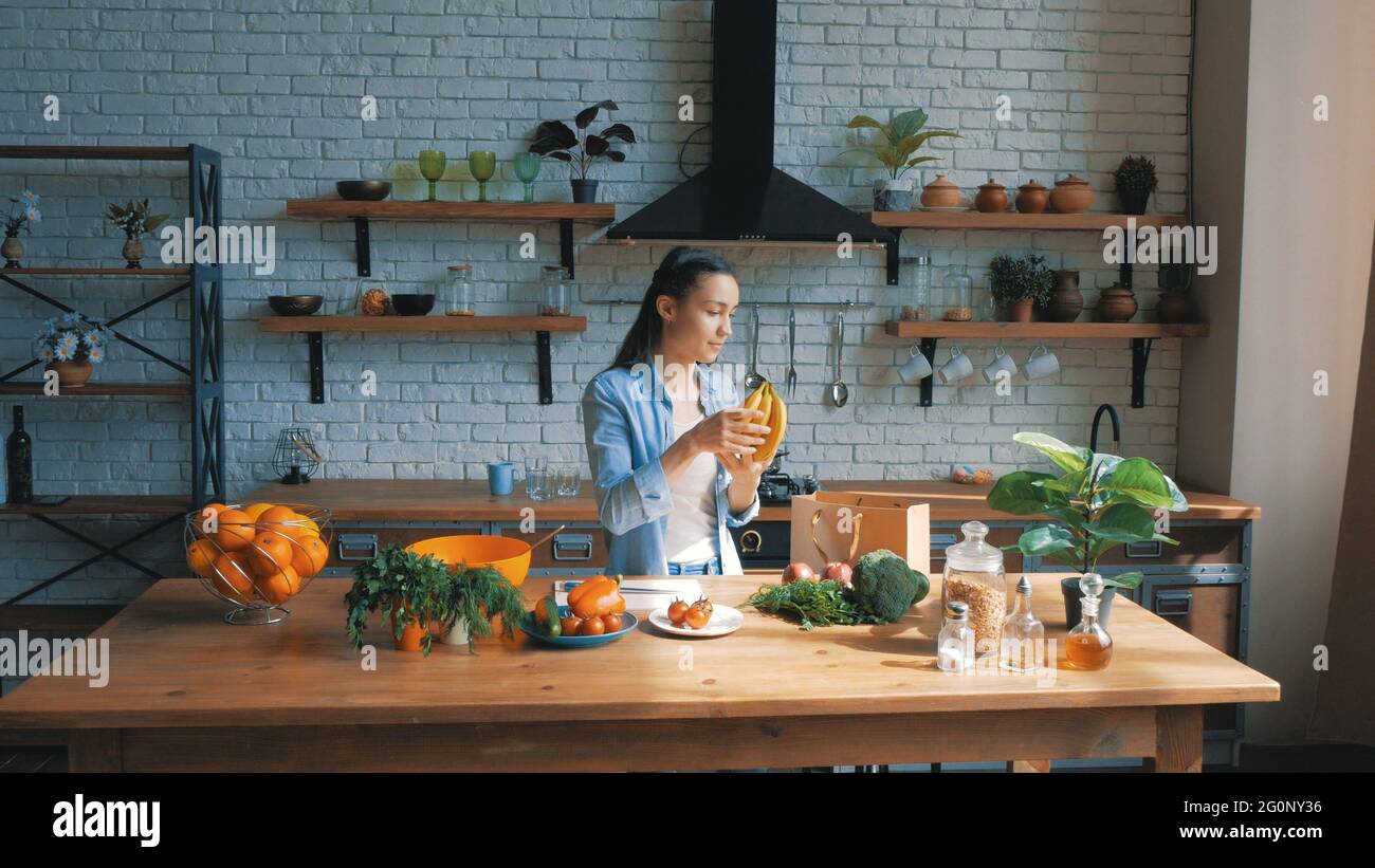 Una bella giovane donna di buon umore sta smistando le verdure da una borsa e metterle sulla tavola della cucina.housewife giovane e felice stabilisce gli acquisti Foto Stock