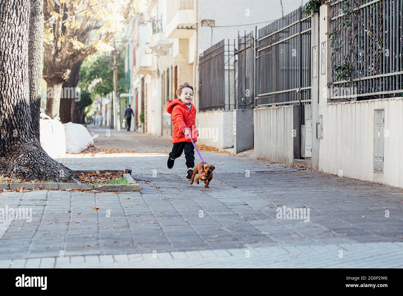 Felice ragazzino che corre con il suo cane dachshund su un marciapiede della città la mattina di un giorno d'autunno. Foto Stock