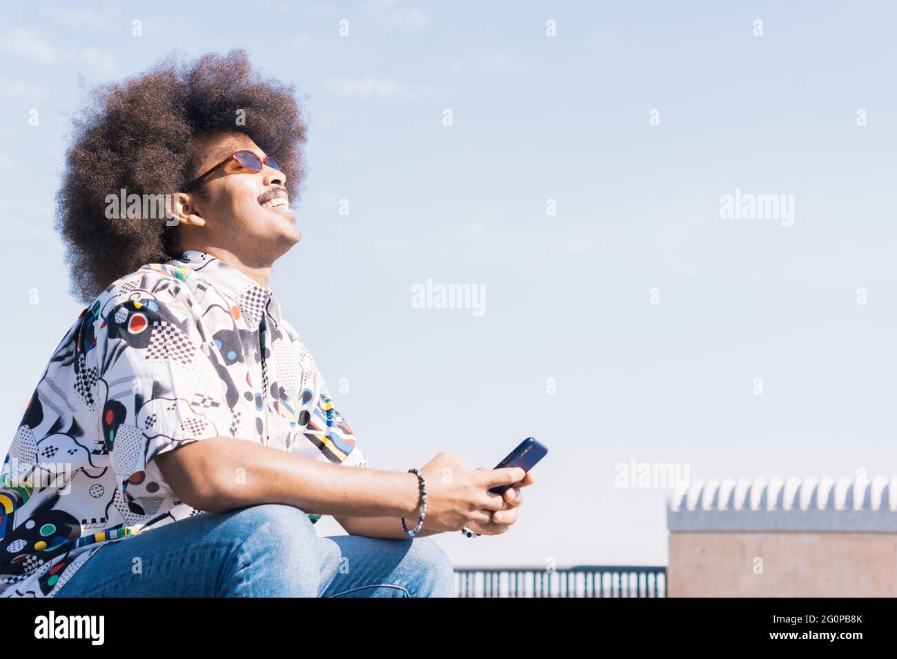 ragazzo nero con capelli afro e occhiali da sole rossi seduti in profilo ridendo con il suo cellulare in mano con il cielo blu sullo sfondo Foto Stock