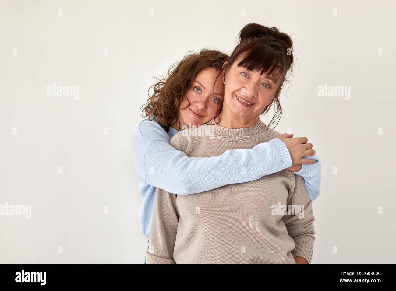 Giovane donna abbracci felice matura sorridente donna che indossa abiti casual stand insieme in camera appartamento con muro beige closeup Foto Stock