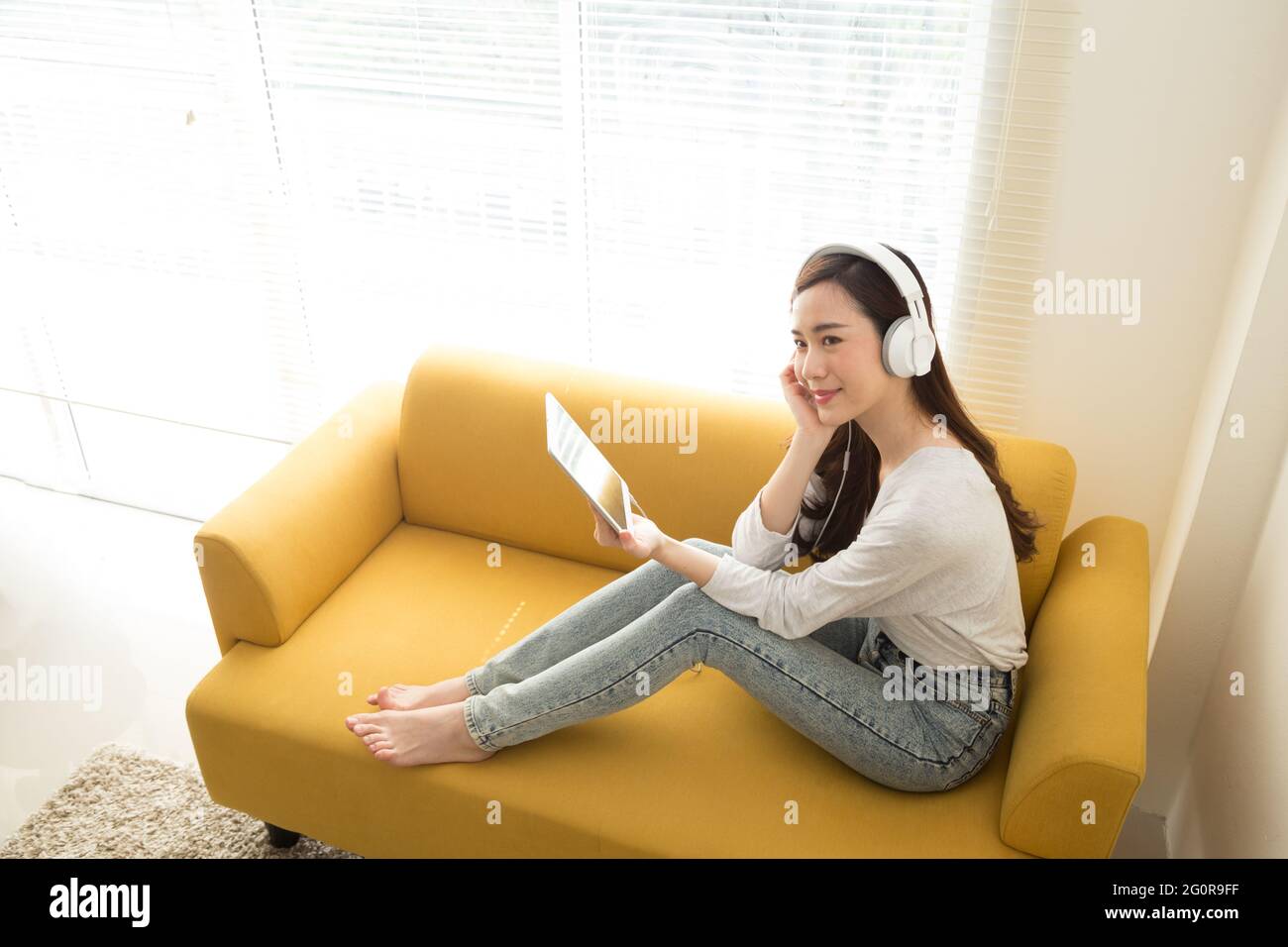 Bella giovane donna asiatica ascolto di musica con cuffie utilizzando un tablet sul divano in camera, adolescente godendo Top chart servizio di streaming musicale in Foto Stock