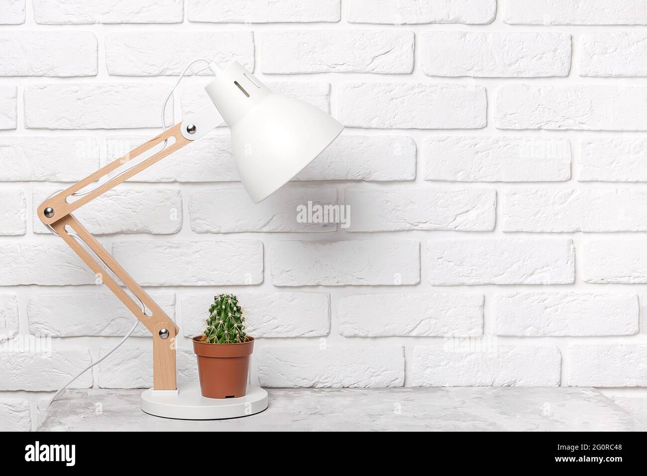 Moderna lampada da tavolo e cactus sul tavolo, sullo sfondo di una parete di mattoni bianchi. Luogo di lavoro, vista frontale, spazio per la copia. Foto Stock