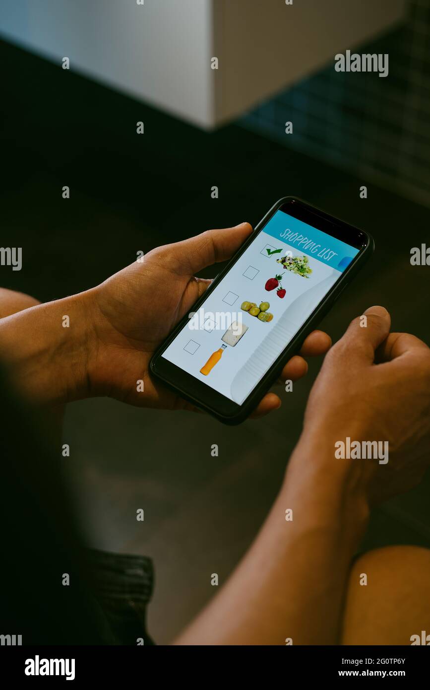 un giovane uomo caucasico, seduto nel gabinetto, sta facendo la sua lista di shopping nel suo smartphone, con una mock app con immagini di diversi prodotti in esso Foto Stock