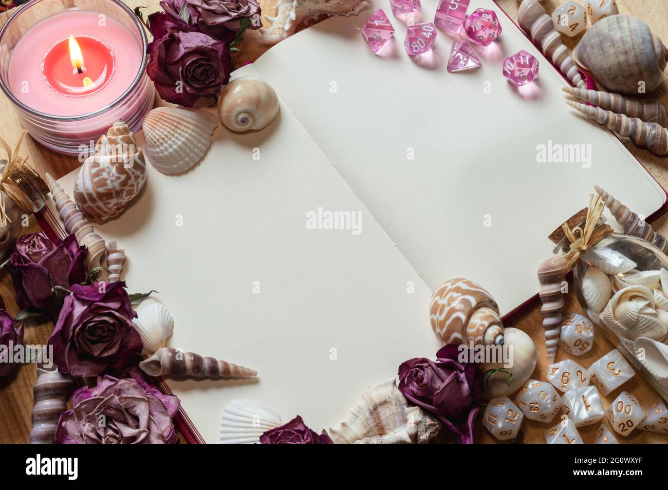 Immagine di un notebook aperto circondato da rose rosa secche, conchiglie, una candela rosa e dadi da gioco RPG bianchi Foto Stock