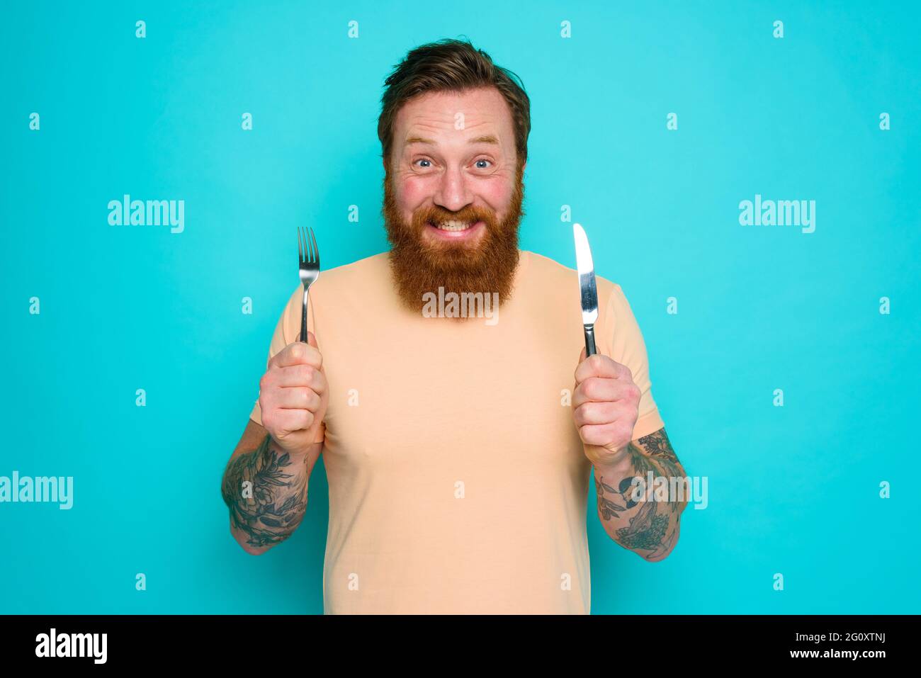 L'uomo felice con i tatuaggi è pronto a mangiare con le posate in mano Foto Stock
