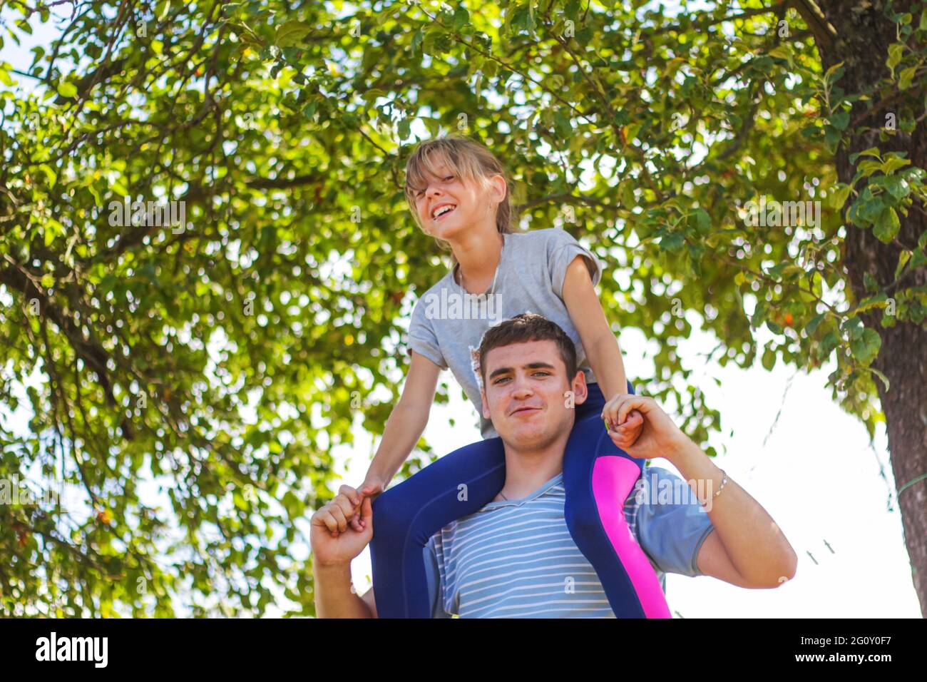 Fratello disinnescato che cavalcava la sorella sulla schiena. Ritratto di ragazza felice sulle spalle dell'uomo, piggyback. La ragazza vola, alza la mano. Famiglia che gioca all'esterno. Albero verde Foto Stock