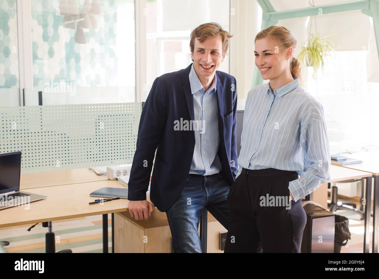 Un paio di colleghi in un nuovo ufficio leggero ridendo. L'uomo e una donna si levano in piedi vicino alle scrivanie nello spazio di lavoro e guardano via Foto Stock