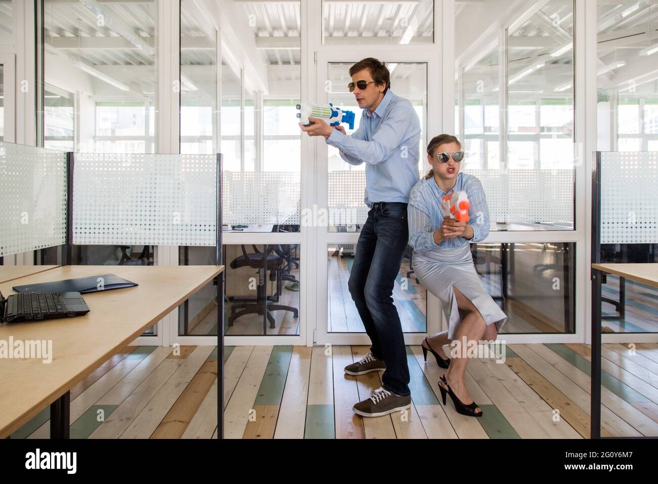 Uomo e donna in abiti formali che si levano in piedi indietro e puntano le pistole giocattolo mentre si divertono in ufficio Foto Stock