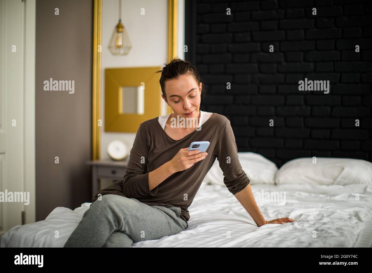 Donna adulta in abiti casual seduta a croce zampe sul letto e smartphone di navigazione in una camera da letto accogliente a casa Foto Stock