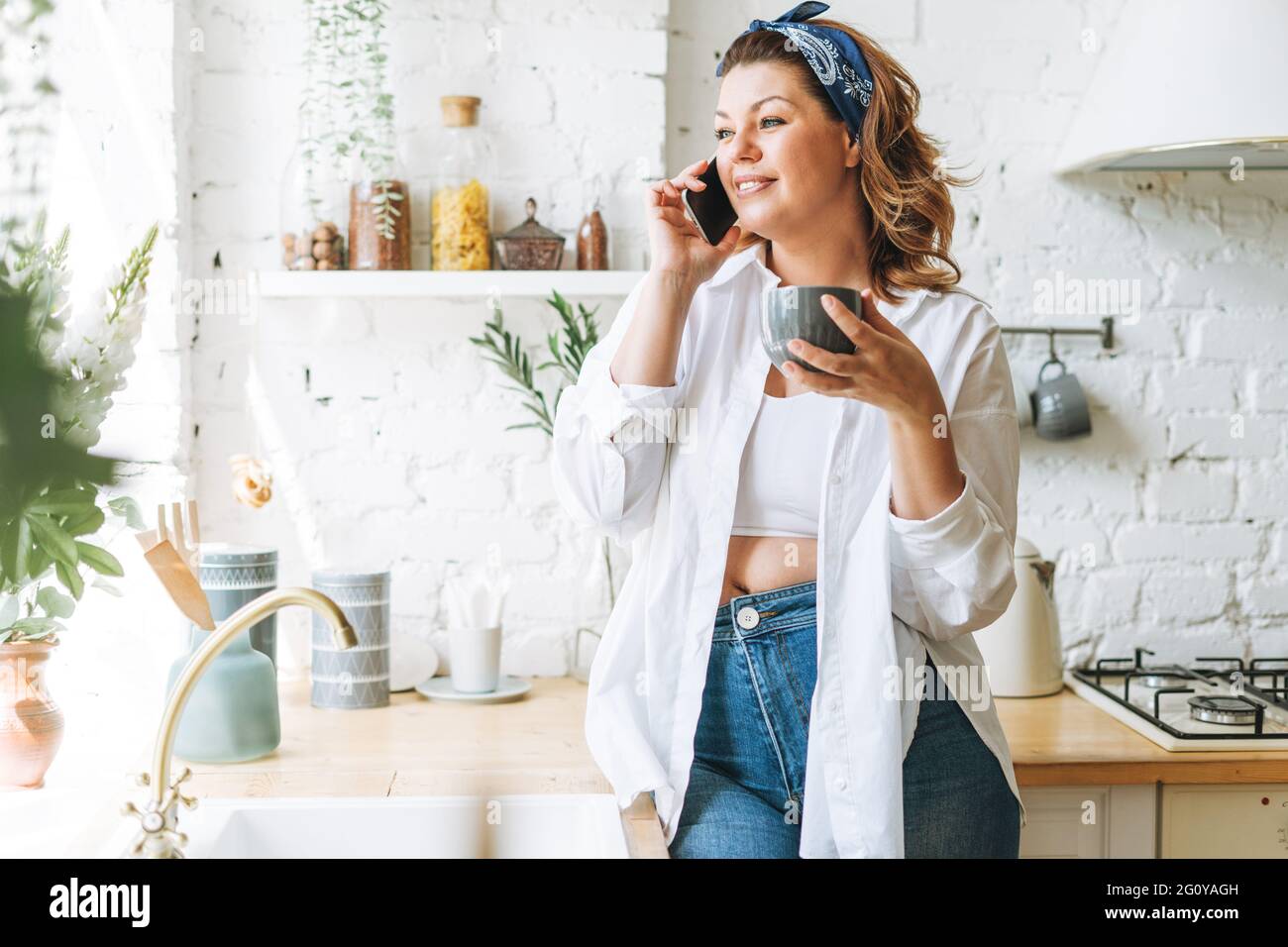 Attraente giovane donna più corpo di taglia positivo in jeans blu e camicia bianca che parla sul cellulare alla cucina domestica Foto Stock