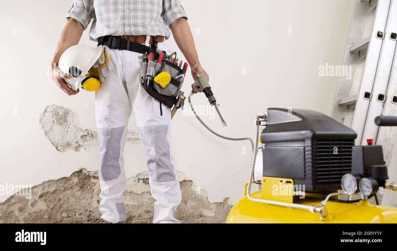 uomo di costruzione che indossa una cintura portautensili, casco con cuffie e scalpello pneumatico a martello pneumatico nelle mani, compressore e scala su bianco Foto Stock