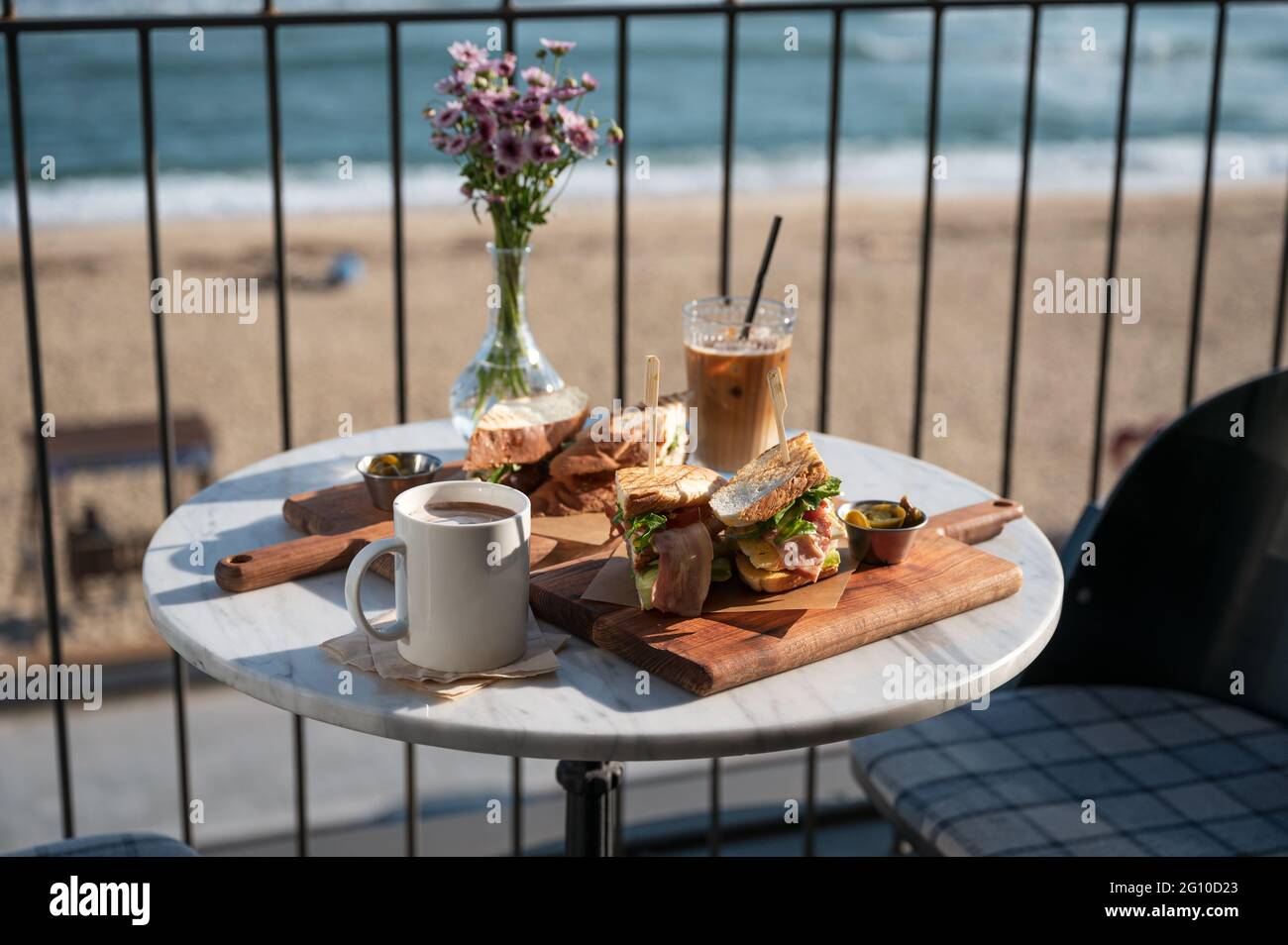 Sandwich di pollo e caffè ghiacciato con cacao caldo e fiore in vaso su tavolo di marmo nel caffè vicino alla spiaggia al mattino Foto Stock
