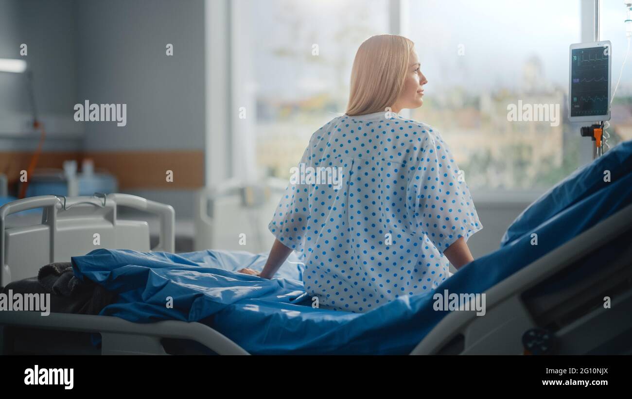 Hospital Ward: Bella paziente caucasico femminile seduta su un letto, completamente in recupero dopo un intervento chirurgico di successo, Covid-19, malattia si guarda intorno e Foto Stock