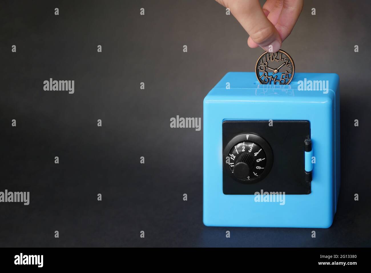 Una mano sta mettendo una moneta a forma di orologio in una banca di piggy blu a forma di cassaforte. Foto Stock