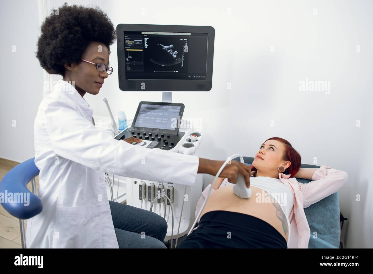 Felice giovane donna caucasica incinta che ha ecografia diagnostica in clinica moderna, guardando la sua donna nera medico ostetrista, mettendo trasduttore a ultrasuoni sul grande cute ventre Foto Stock